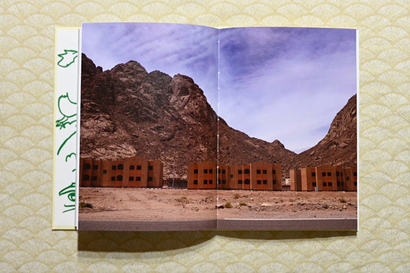Several orange brick buildings feature against a mountainous desert backdrop. Extract from photobook by Rehab Eldalil