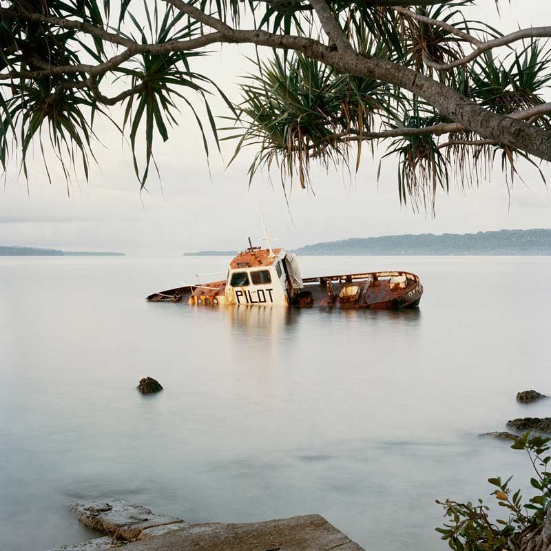 A rusted boat sinks on an angle off a pacific island. The photograph is framed by palm trees and there are islands in the distance. Photo by Jon Tonks