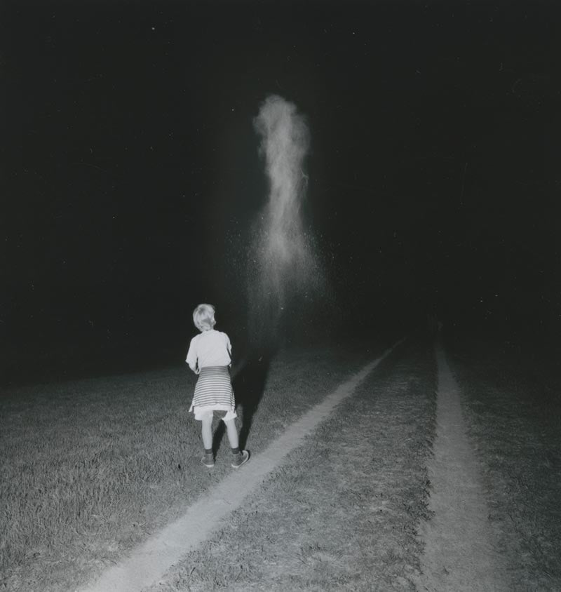 A child stands in a field at night with dirt tracks fading into the distance