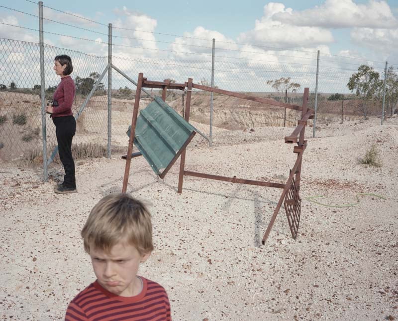 A child and woman stand in a dusty dirt landscape with a barbed wire fence and a rusted metal structure 